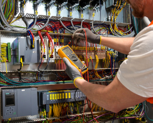 an electrician in a cap, yellow glasses and an orange vest measures electric current with a digital multimeter on a distribution box