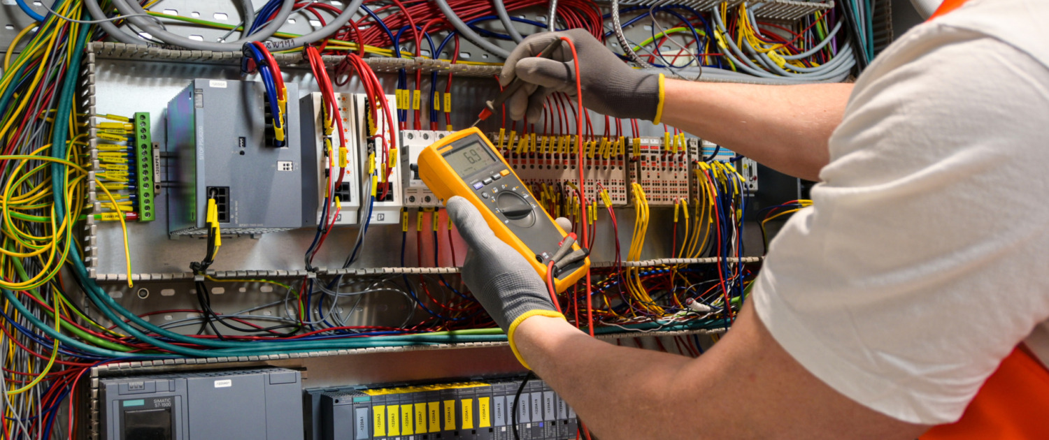 an electrician in a cap, yellow glasses and an orange vest measures electric current with a digital multimeter on a distribution box